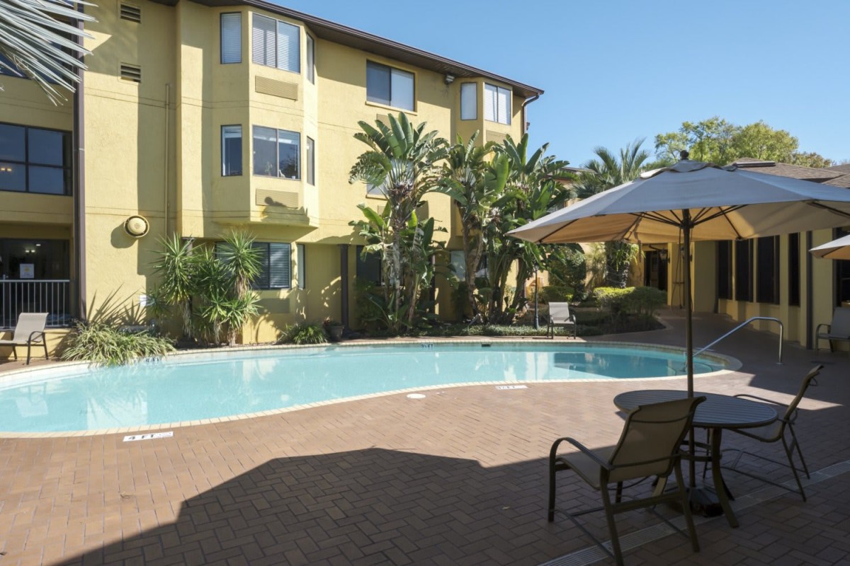 A sunny outdoor pool area surrounded by a yellow apartment building and palm trees.