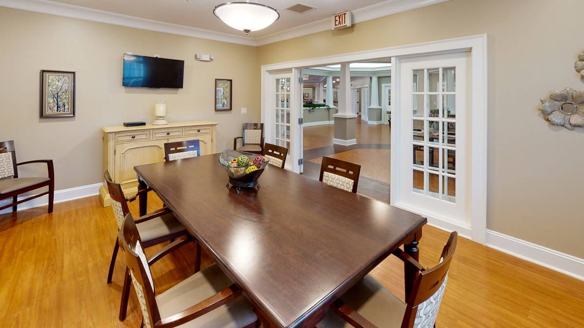 A neatly arranged dining room with a wooden table surrounded by chairs, featuring a wall-mounted television and framed art.