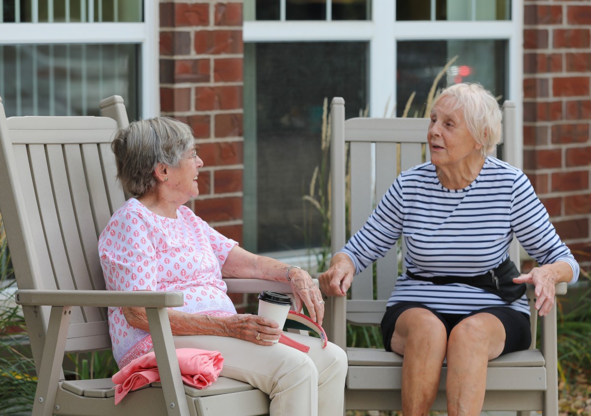 Two elderly women sitting and chatting outside near a brick building.