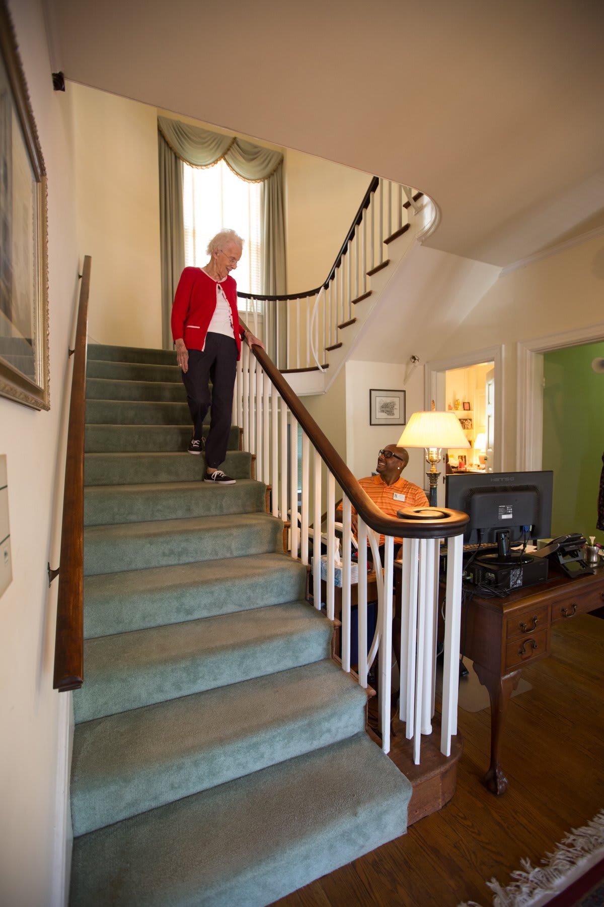 A person in a red jacket descends a staircase while another person sits at a desk in the living room.