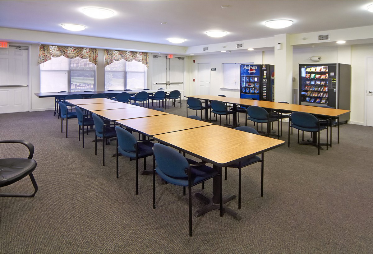 A well-lit conference room with tables arranged in a rectangular formation, surrounded by blue chairs and featuring vending machines along the wall.