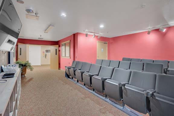 An empty auditorium with rows of seats facing the front, featuring red walls and a carpeted floor.