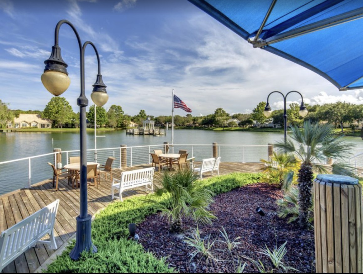 A serene waterfront patio with a garden and seating area overlooking a tranquil lake under a clear blue sky.