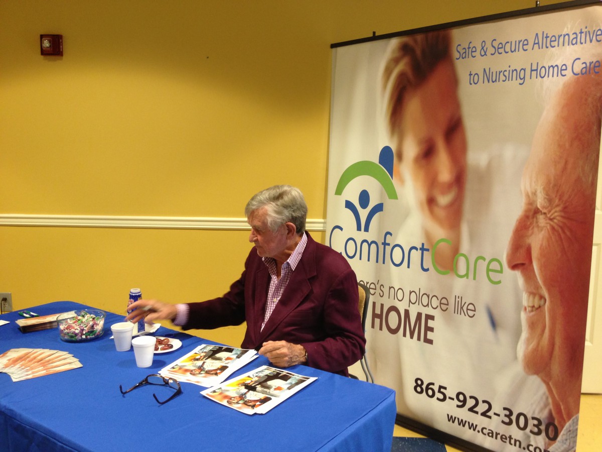 An elderly person is seated at a table during a book signing event, with a healthcare promotional banner in the background.