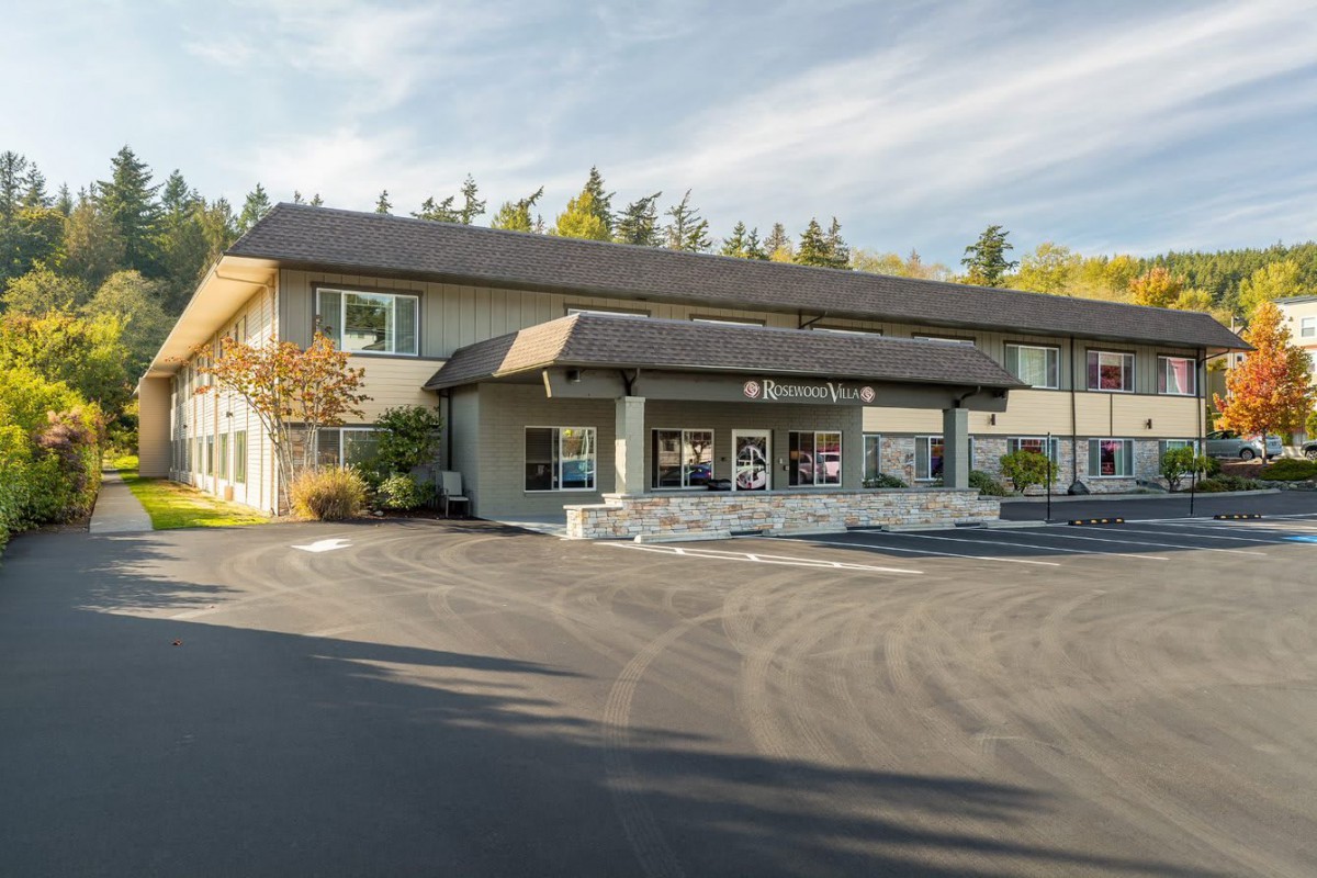 A two-story motel with a parking lot in front, surrounded by trees under a clear sky.