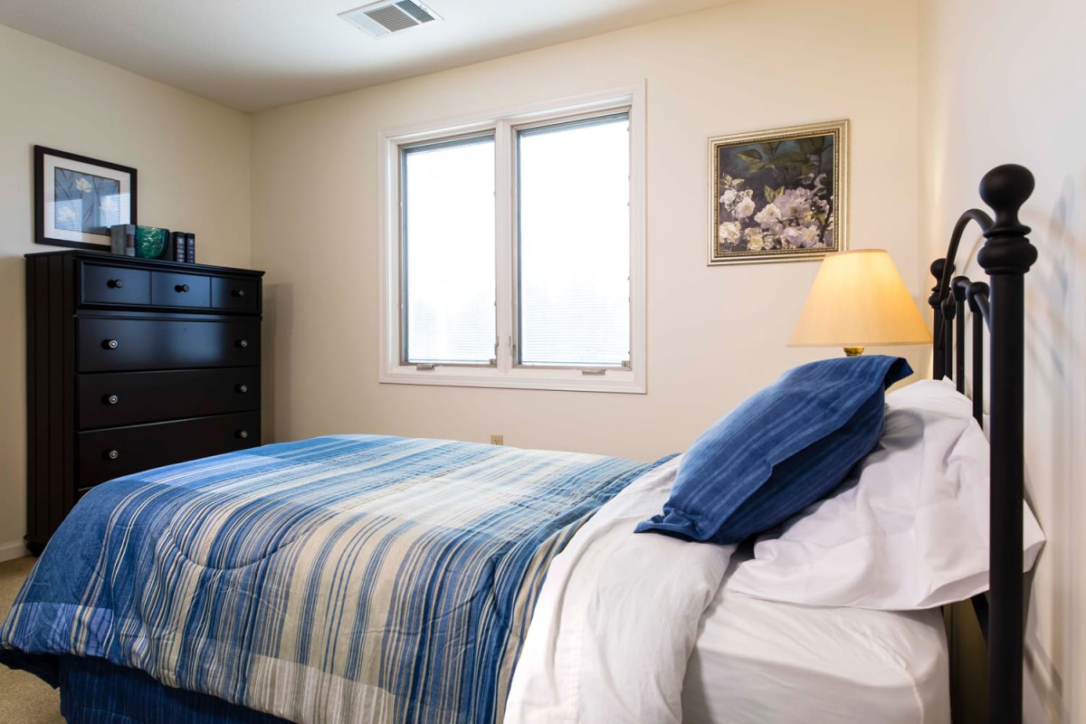 A cozy bedroom with a neatly made bed next to a window, featuring blue and white bedding, a nightstand with a lamp, and additional furniture.