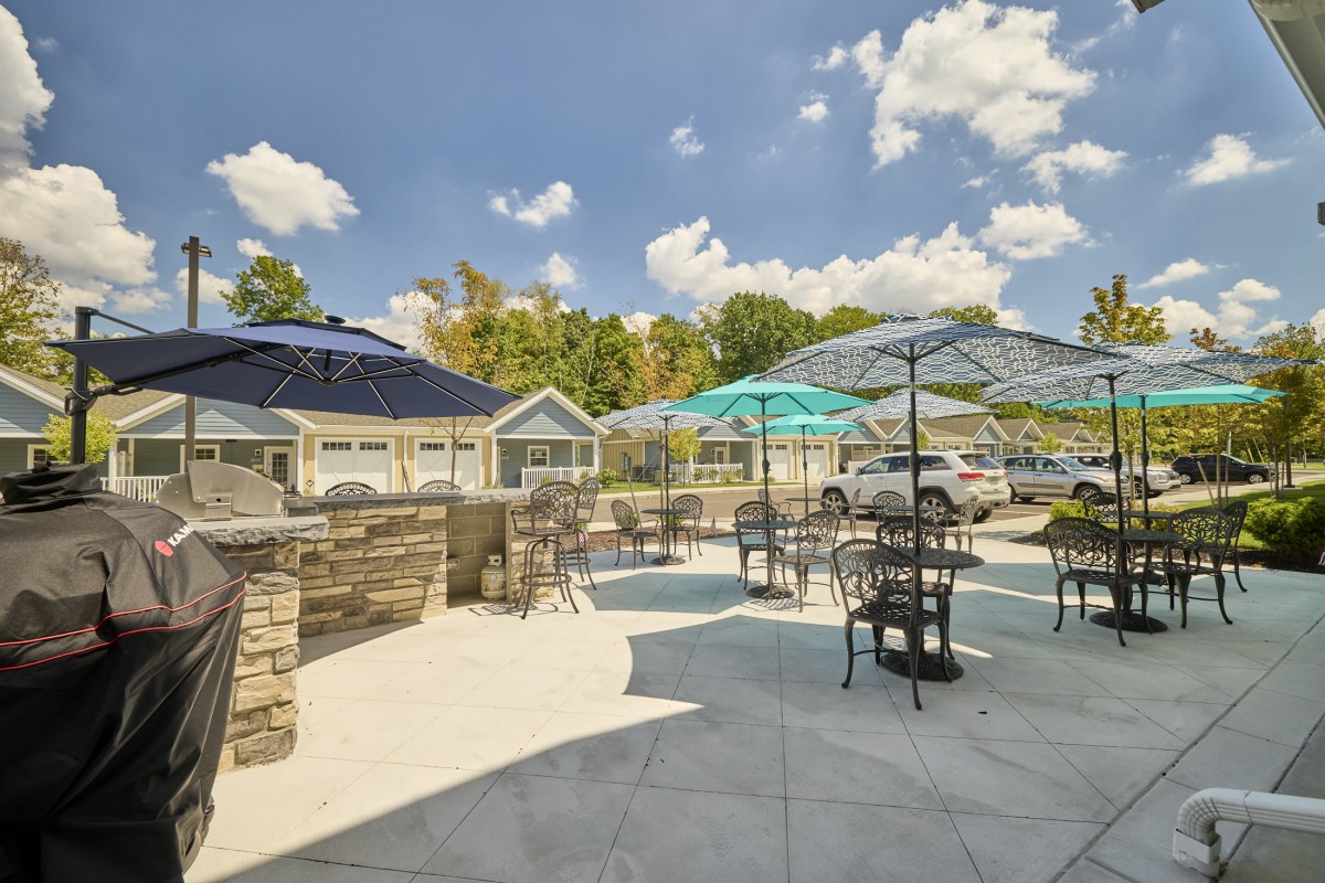 A sunny outdoor patio area with tables, chairs, and umbrellas, surrounded by stone walls and trees.