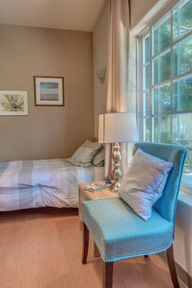 A cozy bedroom with a bed, lamp, and chair by a large window.