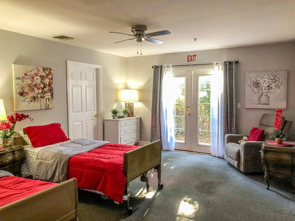 A cozy bedroom with twin beds and bright red bedding, featuring a comfortable chair, lamp, and curtains framing the glass doors.