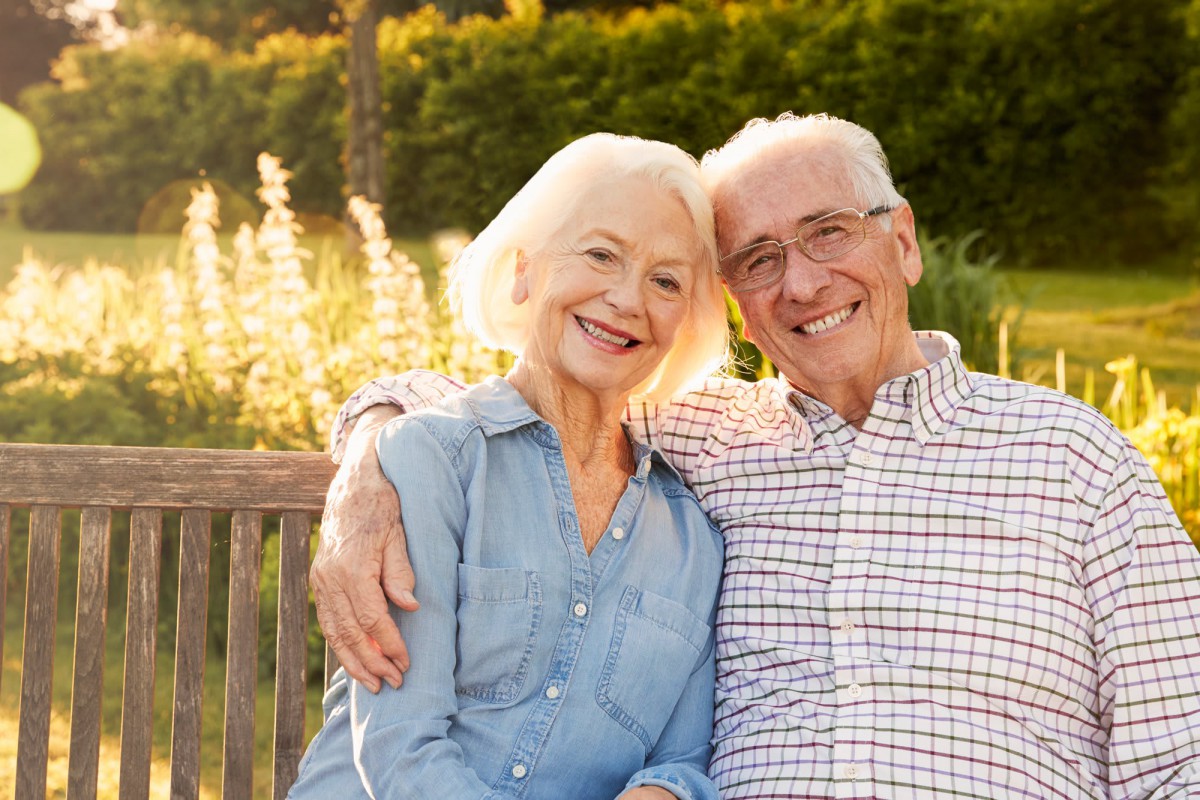 An elderly couple sitting on a bench, smiling and enjoying a sunny day in the garden.