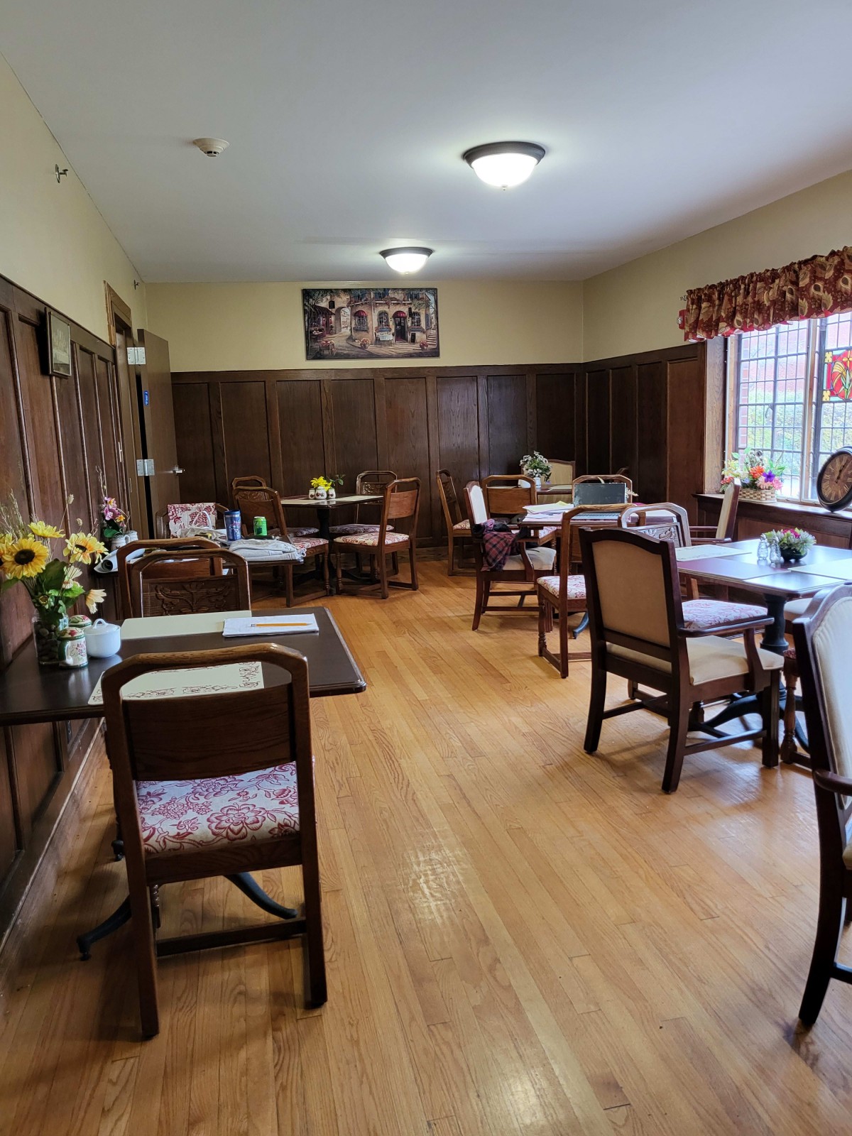 A cozy dining room with wooden floors and several tables set for guests, featuring sunflowers as decoration.