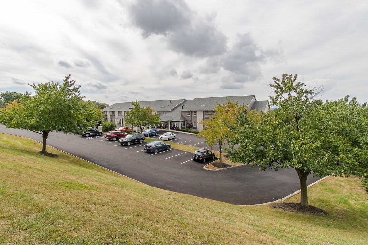 A suburban apartment complex with a large parking lot and surrounding greenery under a cloudy sky.
