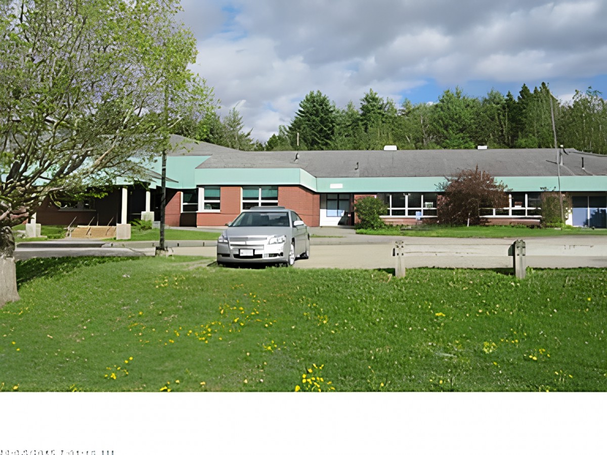 A school building with a car parked in front, surrounded by trees and grass under a cloudy sky.