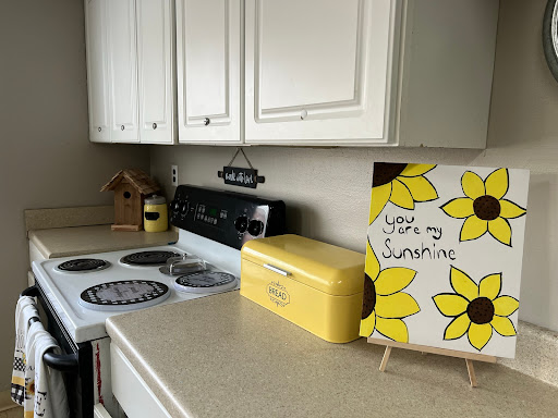A bright and cheerful kitchen corner featuring a sunflower-themed artwork, a yellow breadbox, and an inviting cooking space.