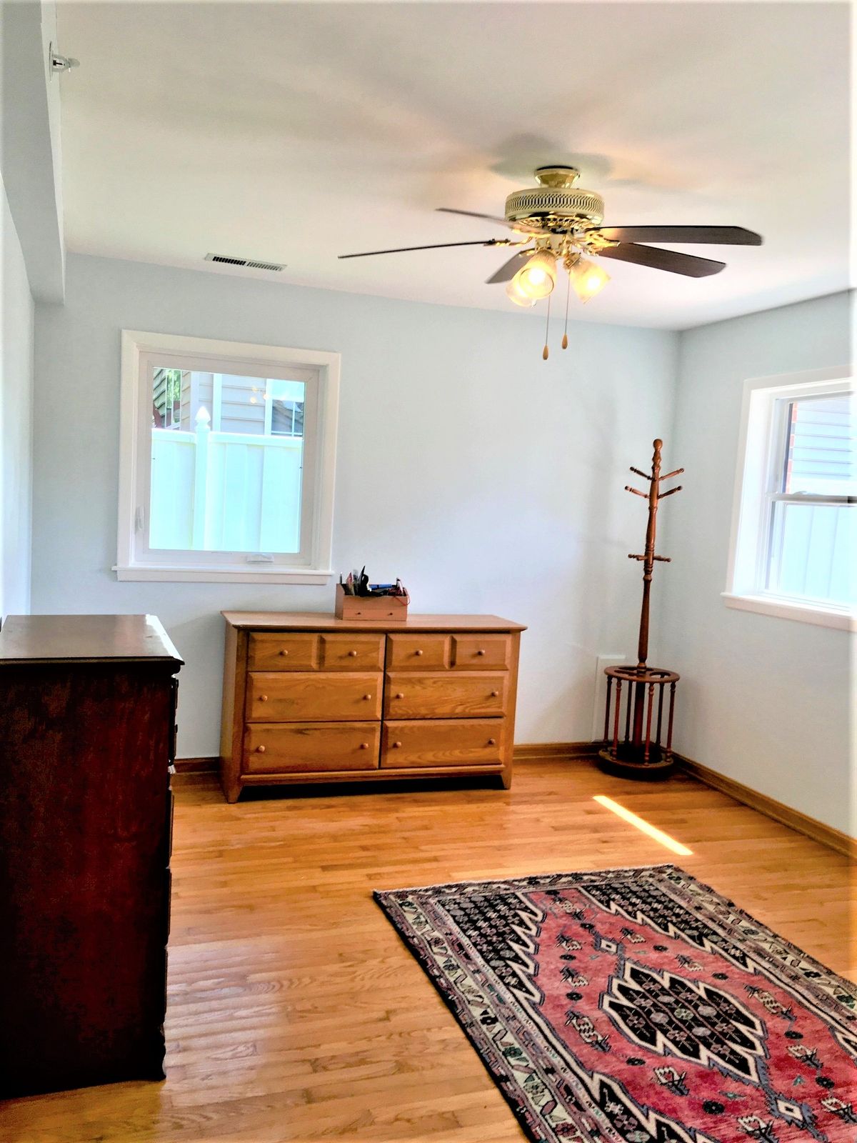 A bright bedroom with wooden floors, featuring a dresser, ceiling fan, and windows allowing natural light.