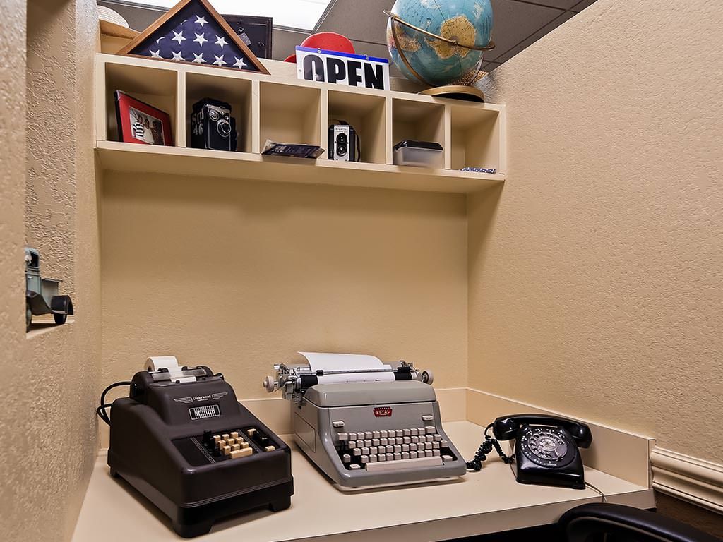 A vintage office setup featuring a typewriter, rotary phone, and calculator on a desk with shelves above.