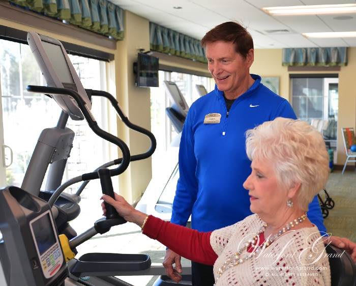 An elderly woman exercising on an elliptical machine with the guidance of a trainer.