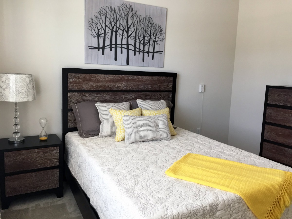 A modern bedroom with a stylish bed, featuring yellow and gray pillows, and a matching blanket. A piece of artwork with trees hangs above the headboard.