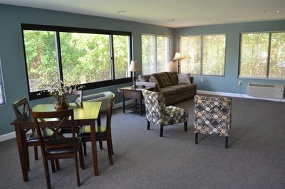 A cozy living room with a sofa, patterned chairs, and a dining table beside large windows.