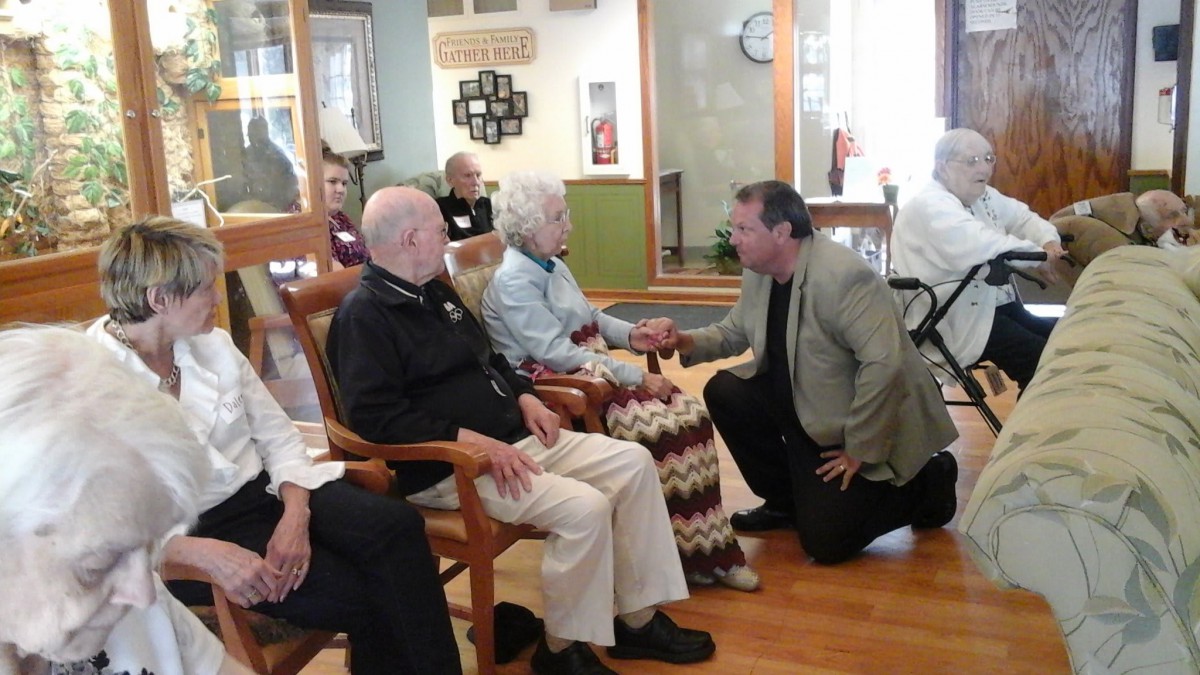 A man kneels to speak with elderly residents at a nursing home.