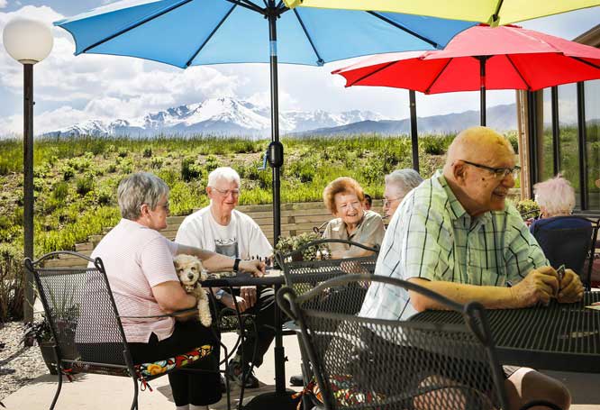 A group of seniors enjoying a gathering on a patio with colorful umbrellas and a scenic mountain view in the background.
