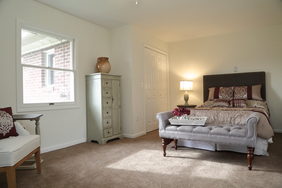 A cozy bedroom with a bed, sofa, cabinet, and a lamp, featuring neutral tones and natural light.
