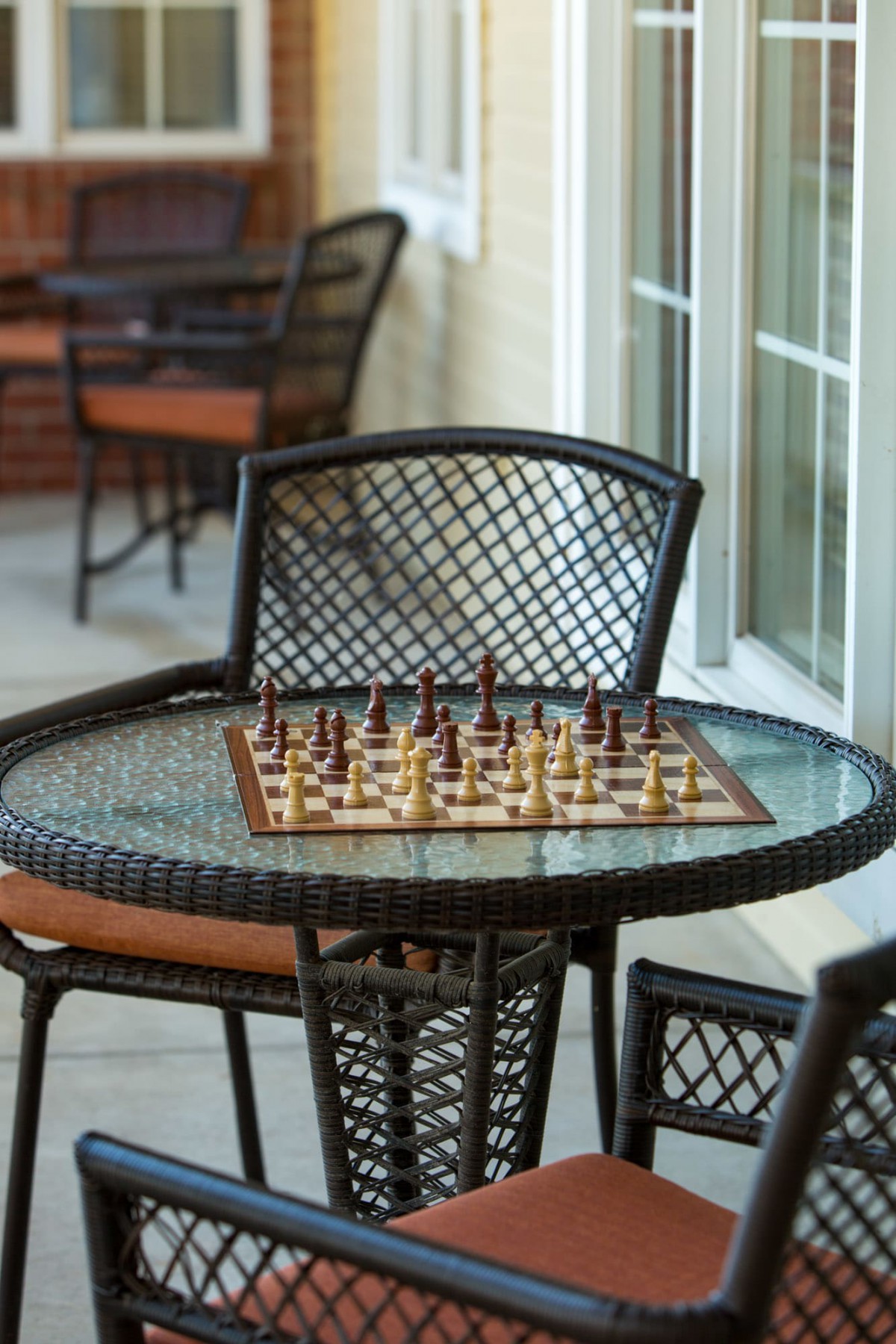 An outdoor chessboard set up on a glass-top patio table with chairs around it.