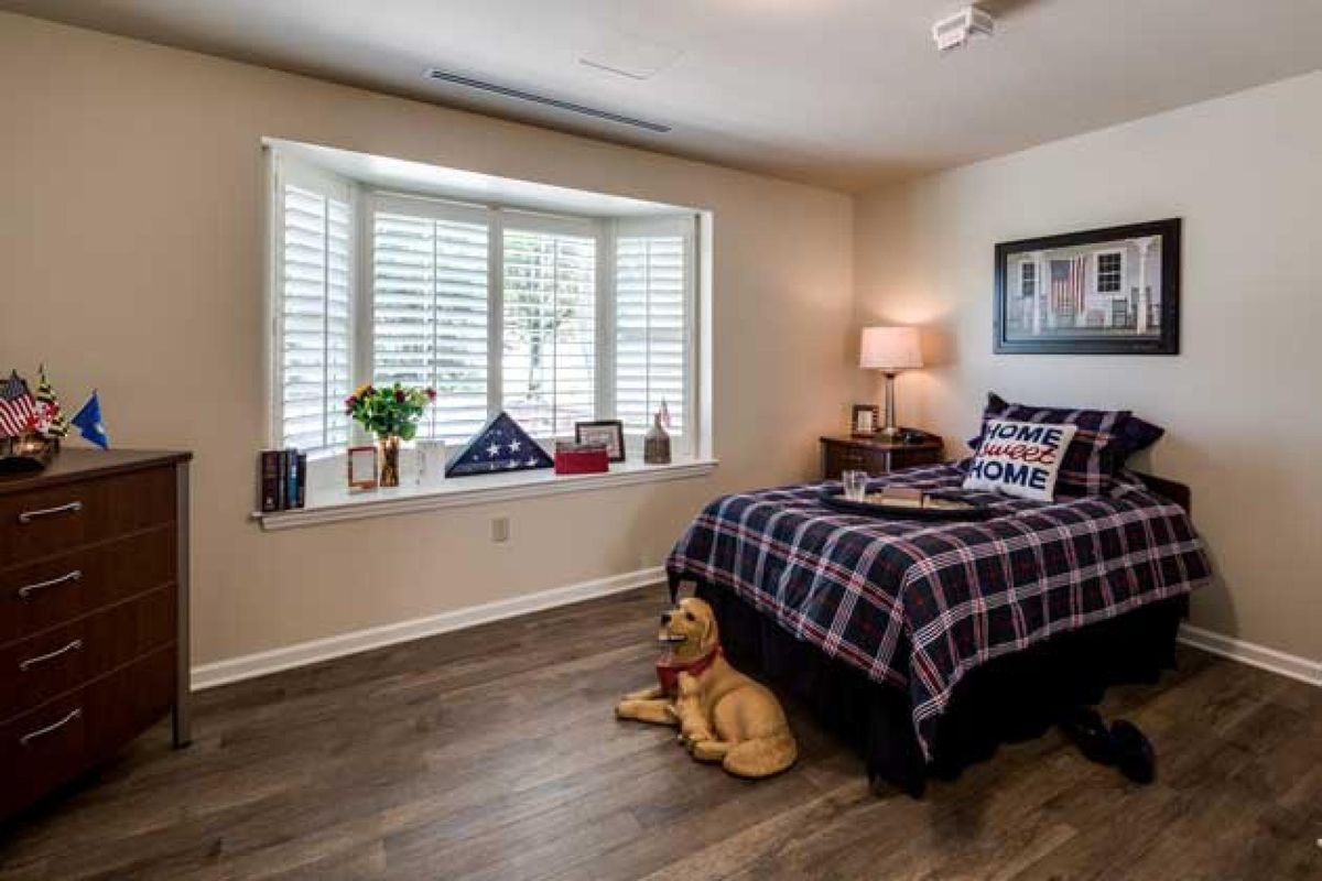 A cozy bedroom with a plaid bedspread, a window seat adorned with decorative items, and a soft stuffed animal on the wooden floor.