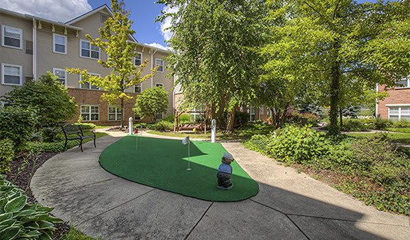 A sunny courtyard featuring a mini golf putting green surrounded by lush greenery and trees.