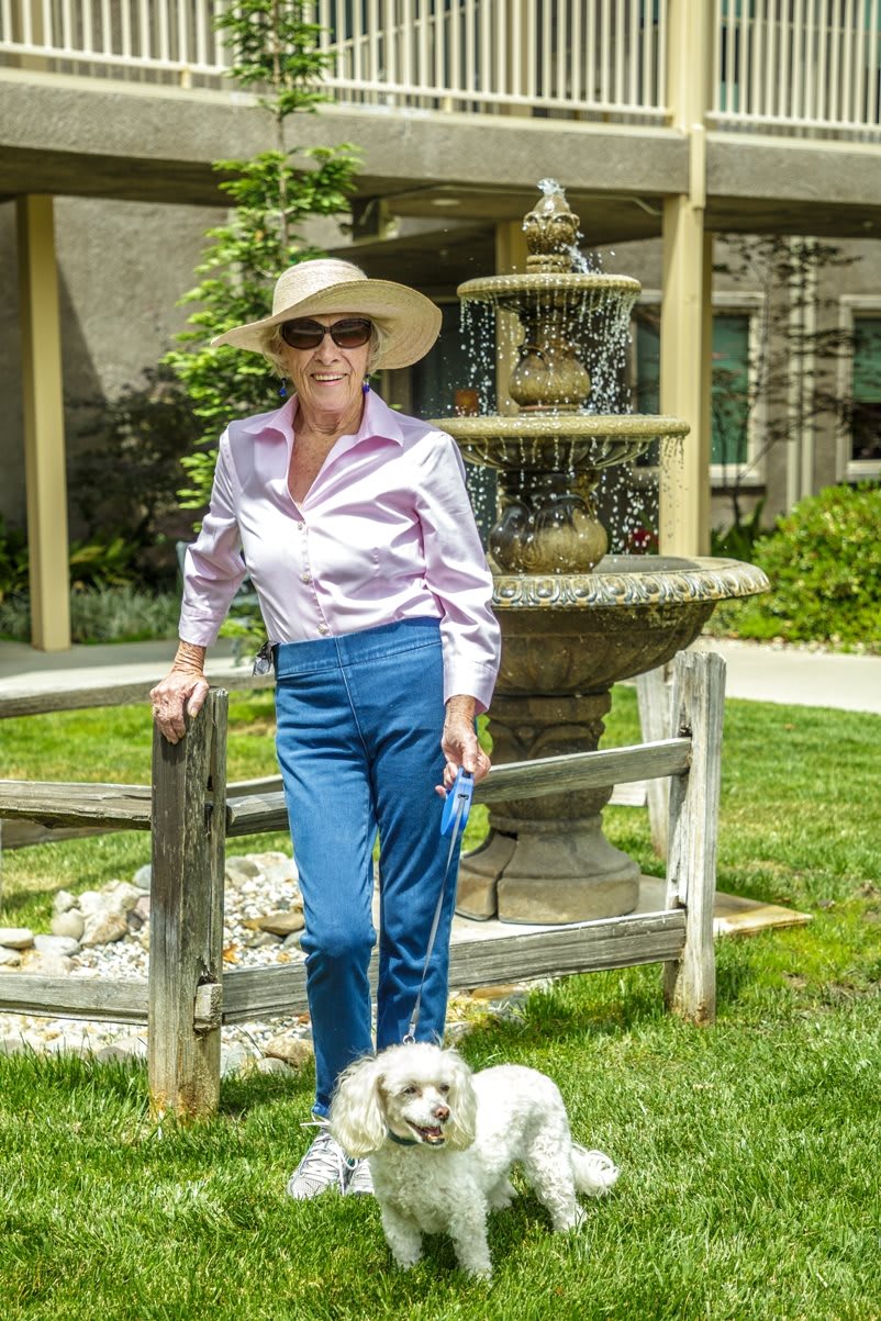 A woman in a hat and sunglasses stands by a garden fountain, accompanied by her dog.