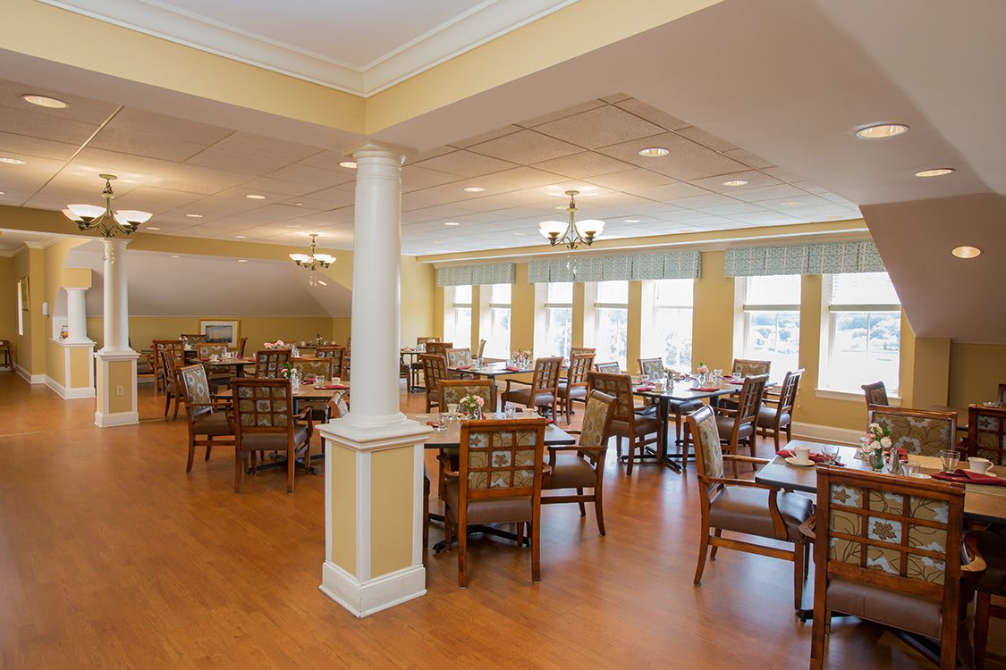 A spacious and well-lit dining room with multiple tables and chairs, featuring large windows and elegant lighting.
