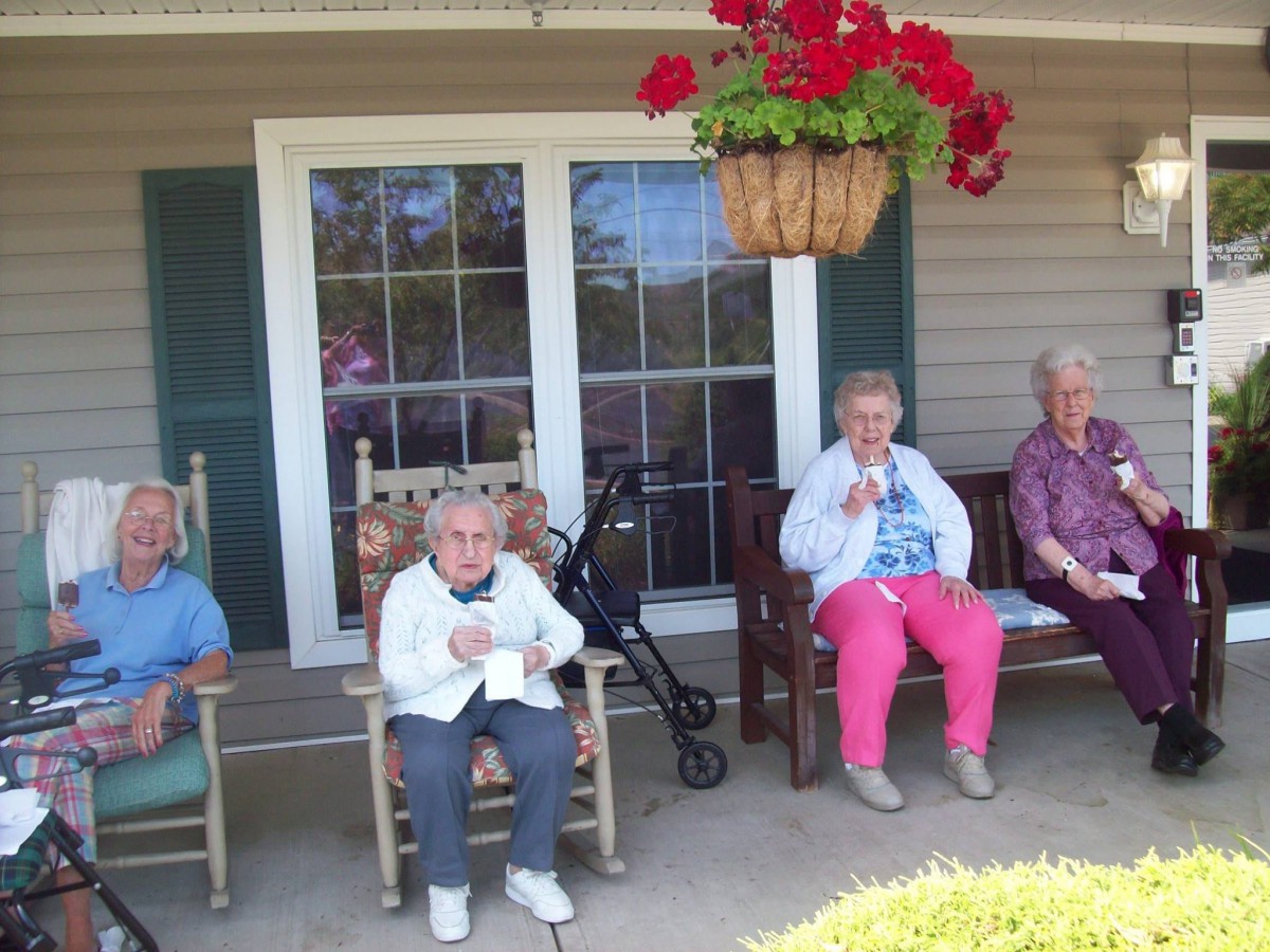 A group of elderly people sitting on a porch in rocking chairs, enjoying the outdoors under a hanging plant.