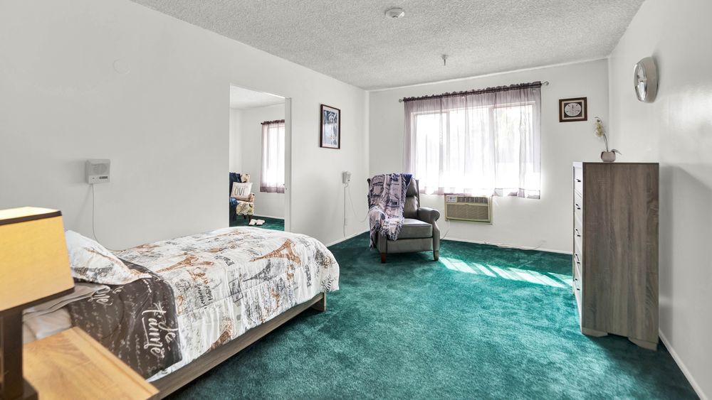A cozy bedroom with green carpet, featuring a bed and a comfortable chair near a sunlit window.