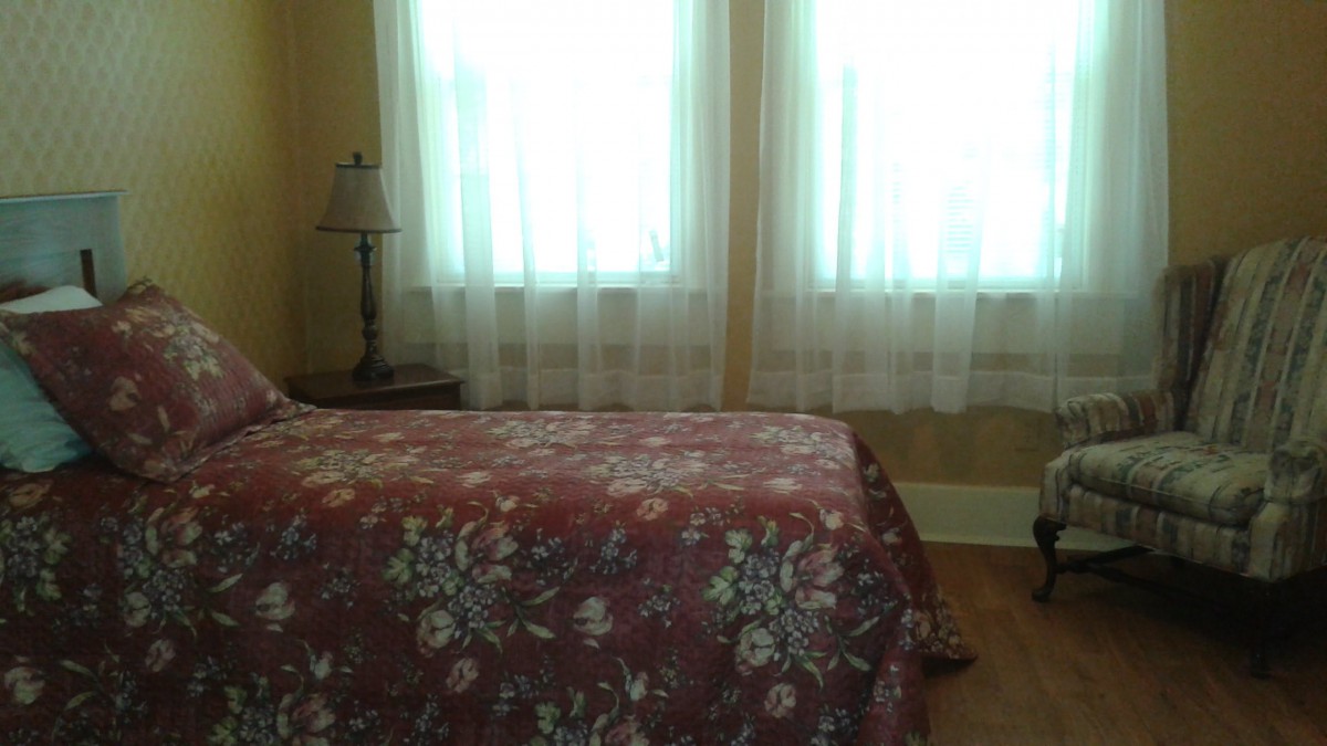 A cozy bedroom with a floral bedspread, bedside lamp, and sheer curtains.