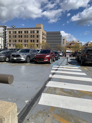 A bustling parking lot with various cars, set against a backdrop of urban buildings and a partly cloudy sky.