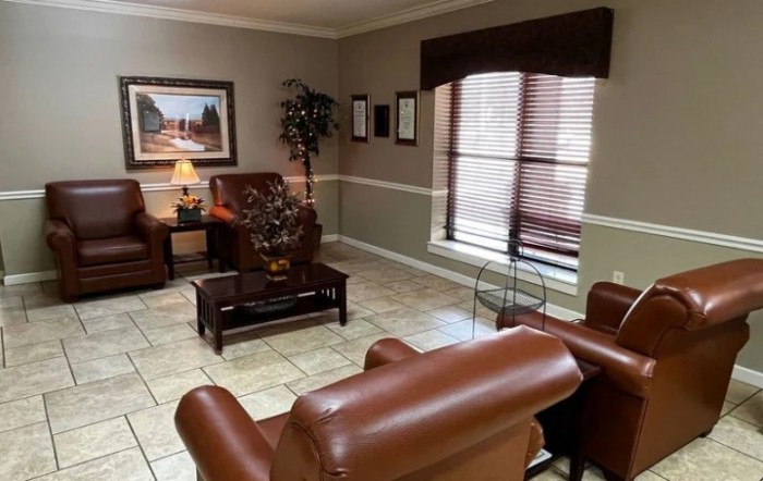 A cozy waiting room with brown leather chairs, a small wooden table, and a decorative lamp under a warm painting.