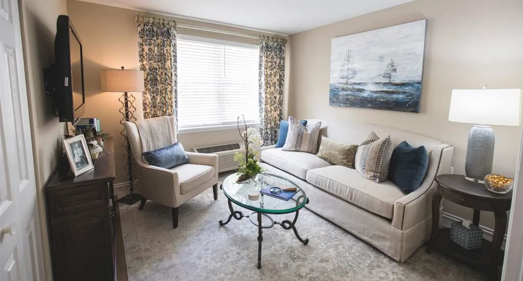A cozy living room with a sofa, armchair, and glass coffee table, decorated with pillows and artwork.