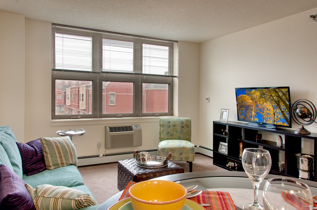 A cozy living room in an apartment with a large window, television, and comfortable seating.