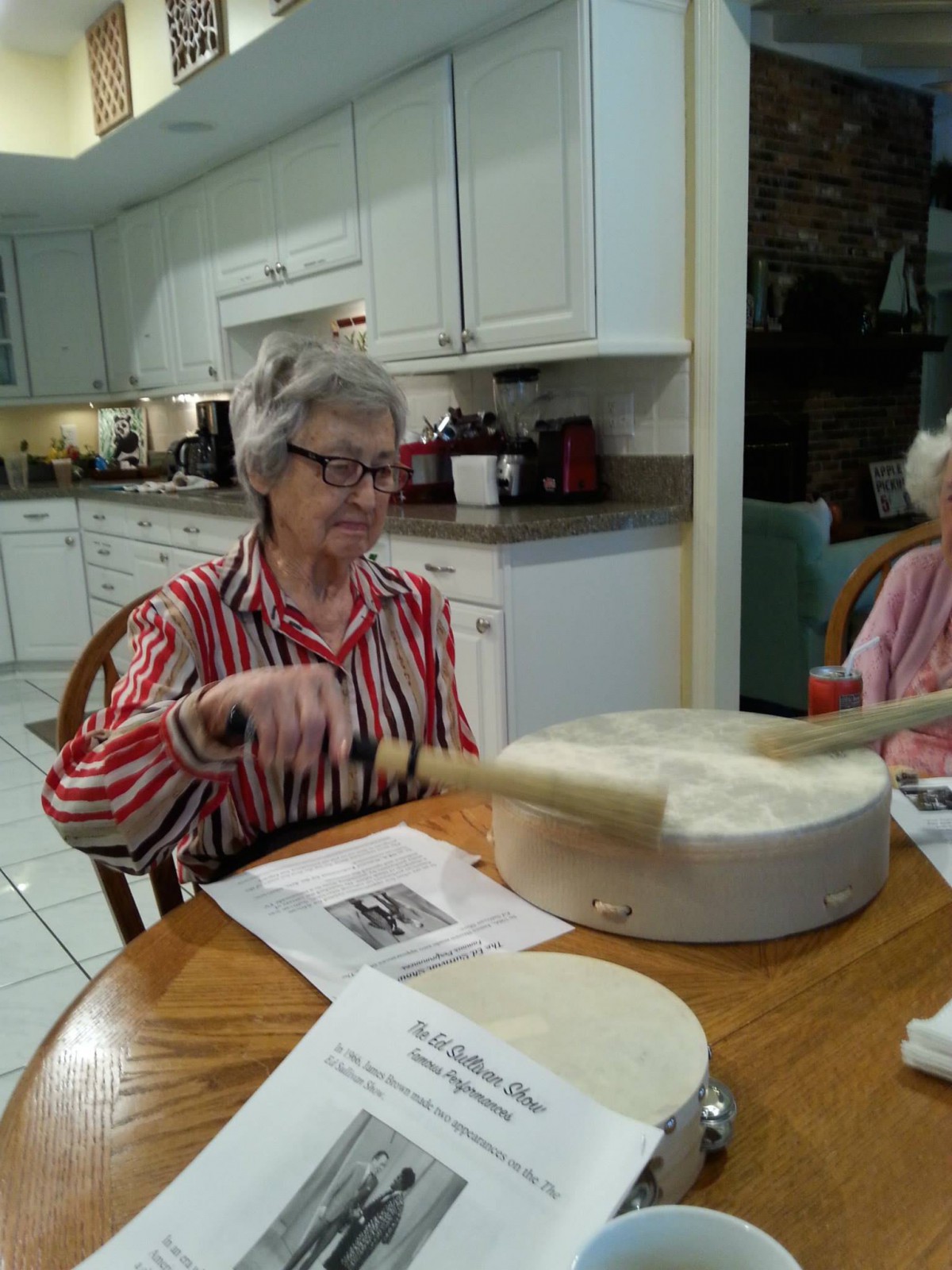 An elderly woman playing a drum in a kitchen setting.