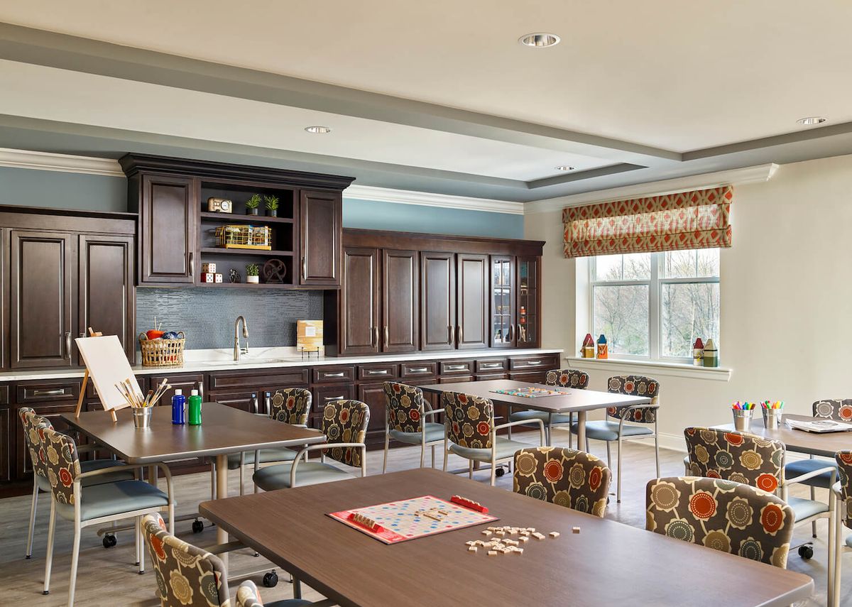 A spacious and well-lit kitchen area with multiple tables and chairs, featuring wooden cabinets and a large window.