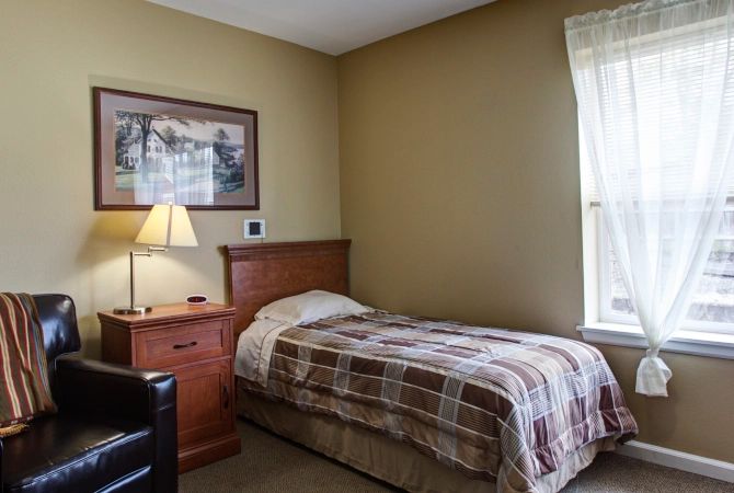 A cozy bedroom with a single bed and a nightstand beside it, illuminated by a table lamp next to a window with curtains.