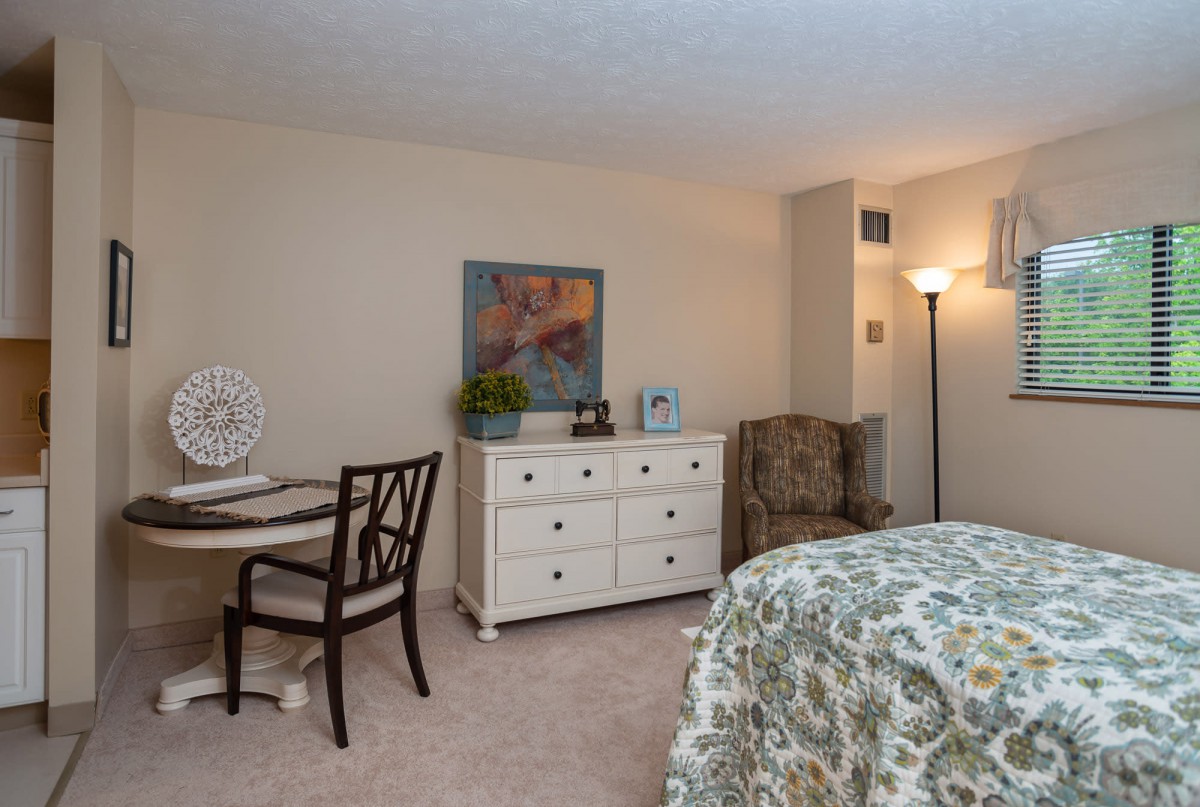 A cozy bedroom featuring a chair, lamp, dresser, and table with decor.