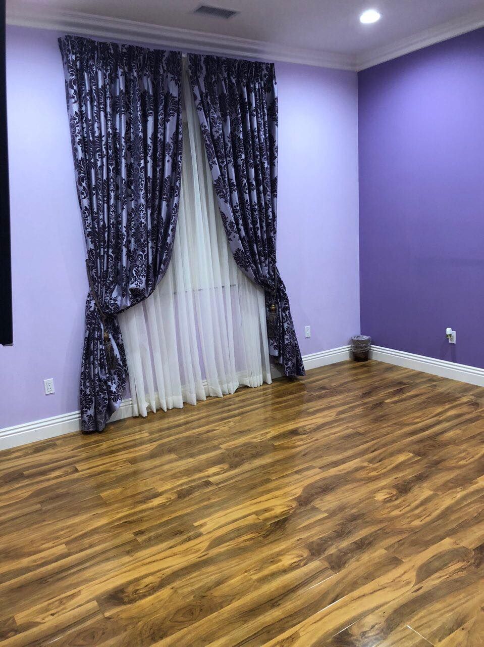 An empty room with wood flooring and purple walls, featuring a window with dark patterned curtains.