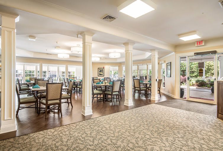 A spacious and well-lit dining area with multiple tables and chairs, surrounded by columns and decorated with carpeting.