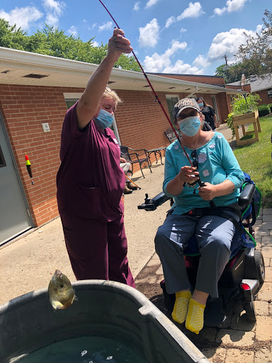A caregiver assisting a person in a wheelchair with fishing on a sunny day.