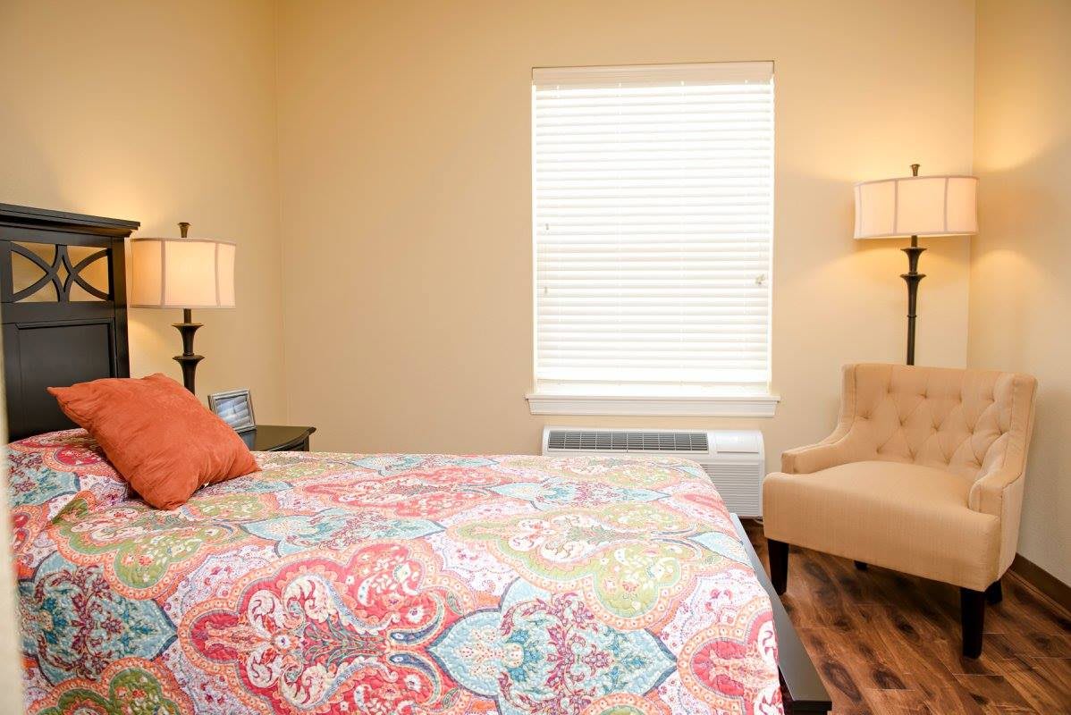 A cozy bedroom with a colorful bedspread, lamps, and a comfortable chair by the window.