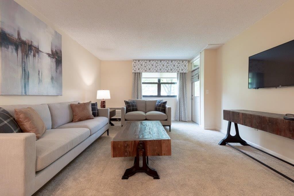 A cozy and well-decorated living room with a comfortable sofa, a wooden coffee table, lamps, and large windows with curtains.