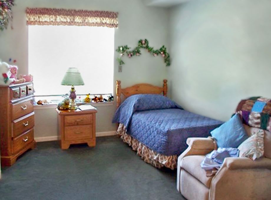 A cozy bedroom with a single bed, nightstand, lamp, and chair next to a window.