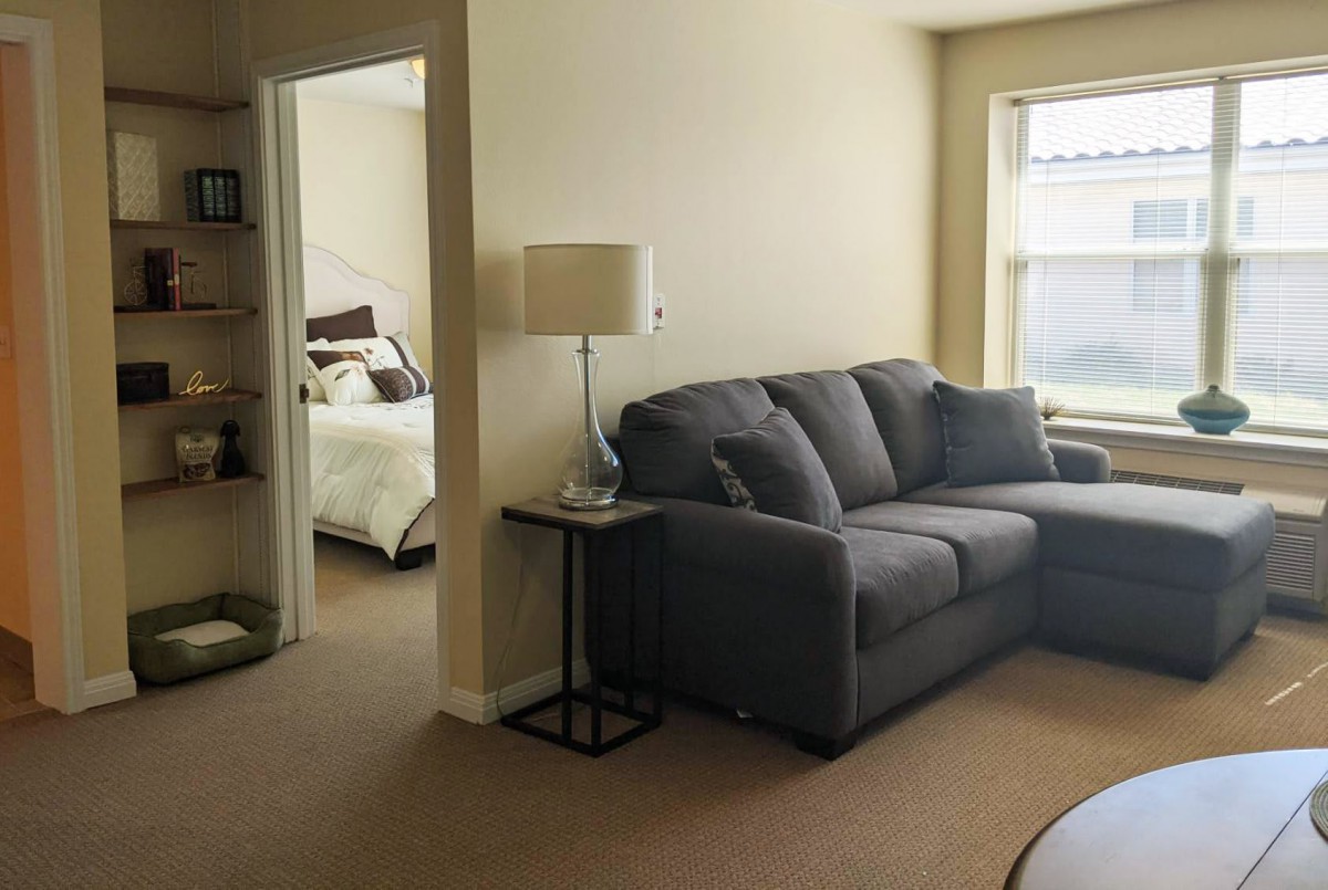 A cozy living room with a grey sofa and a view into a bedroom.