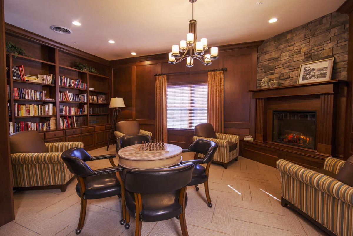A cozy library room with wooden shelves, a fireplace, and a round table surrounded by armchairs.